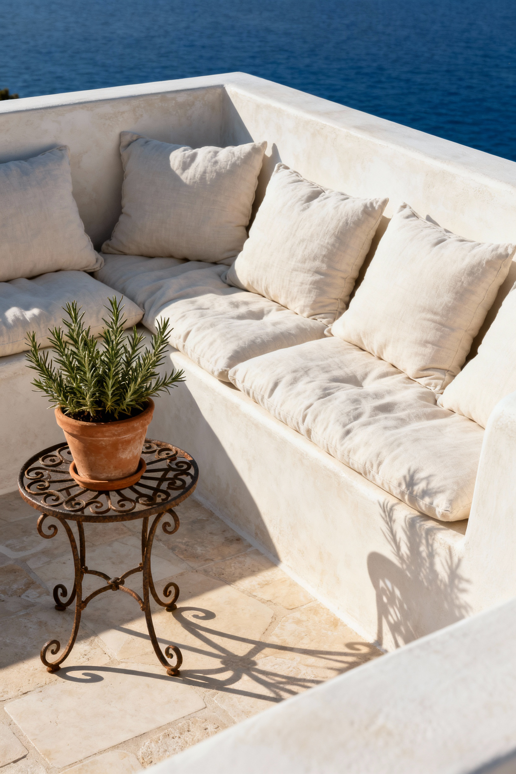 Elegant Mediterranean deck with bespoke built-in banquette, artisan wrought iron table, linen cushions, and rosemary plant, capturing a luxurious seating enclave under morning light.