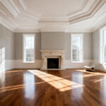Expansive, sunlit living room featuring detailed crown molding, wainscoting, and a limestone fireplace, emphasizing the structural lines and architectural foundation of the space.