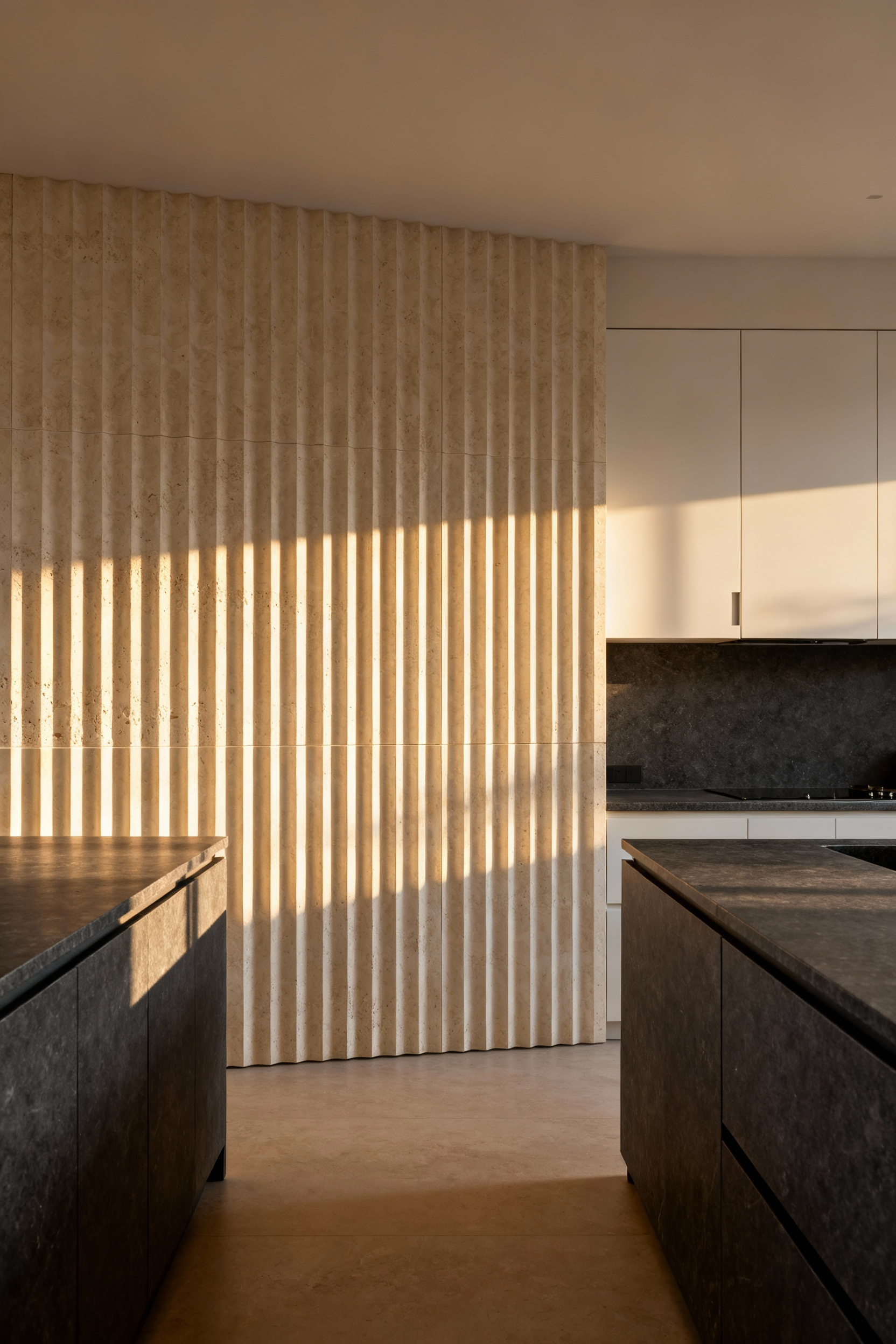 A modern kitchen featuring a floor-to-ceiling fluted limestone backsplash that casts strong, rhythmic shadows from natural directional lighting over contrasting dark gray soapstone countertops.